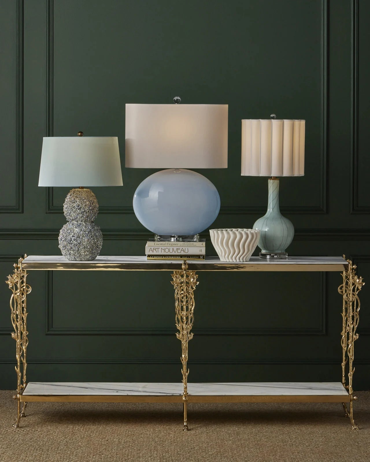 Three designer table lamps on a gold and marble console in front of a dark green wall, featuring the Celadon Crackle Table Lamp by Currey & Company.