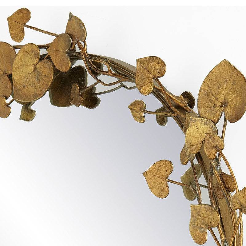 A close up image of the Vinna Brass Round Mirror by Currey & Company with a white background, photo showcasing the intricate details on the leaves surrounding the mirror.  