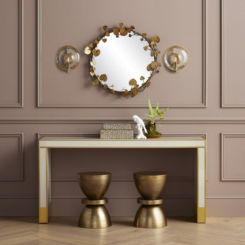 An image of the Vinna Brass Round Mirror by Currey & Company with an antique brass finish, 32 inches in diameter, surrounded by beautiful leaves inspired by nature with a tan wall background set up like an entryway with a console table and two stools.